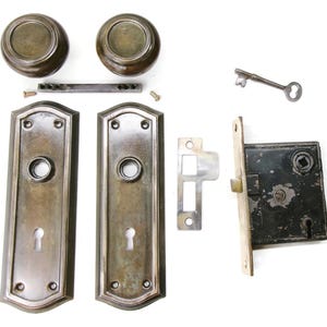 May include: An antique door hardware set, featuring two round doorknobs, two rectangular backplates, a lock mechanism, a strike plate, a key, and small screws. The hardware has a bronze finish and is arranged on a white background.