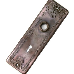 May include: A rectangular, antique bronze door plate with ornate floral and scrollwork designs. The plate features a keyhole and a circular opening for the doorknob. The edges are slightly rounded, and the overall finish has a vintage, aged appearance.