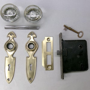 May include: An antique door hardware set featuring two clear glass doorknobs, a metal spindle, brass escutcheons, a strike plate, a black door lock, and a small brass key. The set is designed for a vintage door.