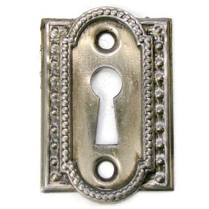 May include: A vintage silver-toned keyhole cover with an oval center and rectangular frame. The keyhole is surrounded by a beaded border and two circular holes. The metal has a distressed finish, suggesting an antique aesthetic.