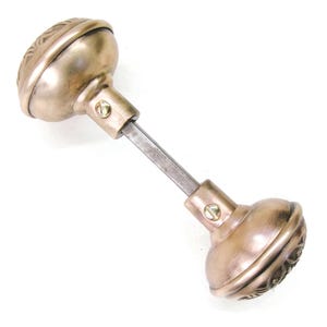 May include: Antique bronze door knob set with decorative floral detailing. The set includes two rounded knobs connected by a metal rod. Each knob is secured with a small screw. The knobs have a warm, metallic finish.