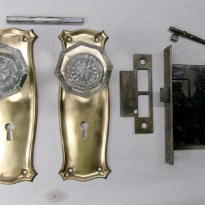 May include: Antique door hardware set featuring two brass door plates with clear glass octagonal doorknobs, a lock mechanism, a strike plate, a key, and a connecting rod. The hardware is aged and shows signs of wear.