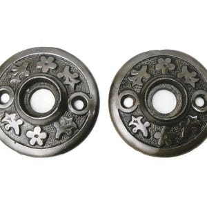 May include: Two antique, dark metal door rosettes with floral and leaf designs. Each rosette has a central hole and two smaller holes for screws. The decorative elements are raised, creating a textured surface.