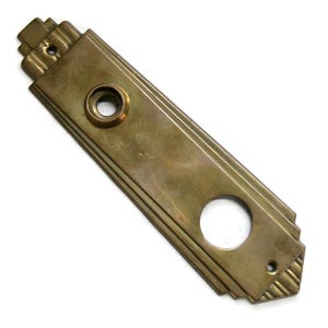 May include: A vintage brass door plate with an Art Deco design. The rectangular plate has a series of parallel lines and a decorative top and bottom. It features a circular hole for a doorknob and a smaller hole for a screw.