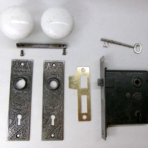 May include: An antique door hardware set featuring two white porcelain doorknobs, ornate metal plates, a key, and a black lock mechanism. The set includes screws and a brass-colored strike plate. Ideal for restoration projects.