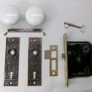 May include: An antique door hardware set featuring two white porcelain doorknobs, ornate bronze-colored faceplates, a black lock, a key, and various small metal pieces. The set is designed for a vintage door.