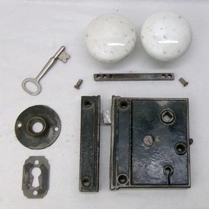 May include: An antique door hardware set, featuring two white speckled ceramic door knobs, a key, a keyhole cover, a door plate, a latch, and screws. The set is made of metal and ceramic.