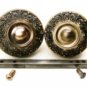 May include: Two antique bronze-colored cabinet knobs with ornate scrollwork around the edges. A metal bar and two screws are also present. The knobs have a circular design with a central, slightly recessed area.