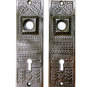 May include: Two antique, rectangular door plates with intricate, embossed floral and geometric patterns. Each plate features a keyhole, a circular opening, and a decorative top with a small hole for mounting. The plates have a dark, metallic finish.