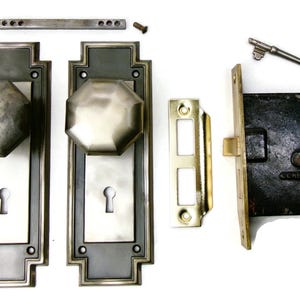 May include: Antique door hardware set, including two octagonal doorknobs, escutcheons, a strike plate, a latch, a key, and screws. The set features a silver and dark gray color scheme, with a vintage aesthetic.