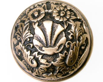 May include: Bronze-toned, ornate cabinet knob with a seashell design at the center, surrounded by floral and foliate patterns. The knob has a detailed, textured surface and a rounded shape, suitable for furniture restoration or decorative projects.