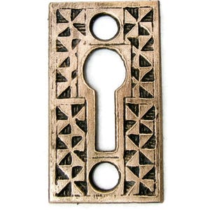 May include: Bronze-colored keyhole cover with a rectangular shape. The cover features a keyhole in the center, two circular holes, and a decorative pattern of triangles and crosses around the edges. The background is white.