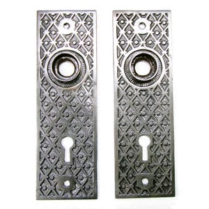 May include: Two antique silver-toned door plates with intricate geometric patterns. Each plate features a keyhole, a circular knob area, and a decorative design. The plates are rectangular and have a vintage aesthetic.