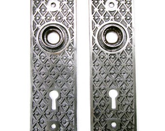 May include: Two antique silver-toned door plates with intricate geometric patterns. Each plate features a keyhole, a circular knob area, and a decorative design. The plates are rectangular and have a vintage aesthetic.