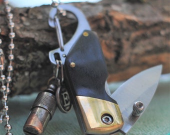 May include: A black and silver pocket knife with a gold-colored handle and a small silver whistle attached to it. The knife is attached to a silver chain.