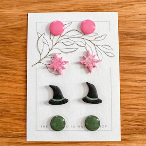 Op de afbeelding: Een set van zes paar oorbellen met verschillende ontwerpen. De oorbellen zijn gemaakt van polymer clay en zijn verkrijgbaar in roze, groen en zwart. De ontwerpen omvatten sterren, heksenhoeden en effen cirkels.