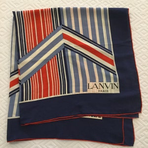 Könnte beinhalten: Ein gefalteter Seidenschal mit einem geometrischen Muster aus roten, weißen und blauen vertikalen Streifen. Der Schal hat einen marineblauen Rand mit roter Paspel. Der Markenname "LANVIN PARIS" ist in Schwarz aufgedruckt.