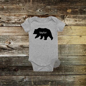 May include: A gray baby onesie with a black bear silhouette and the name "Taylor" printed in white. The onesie has short sleeves and a snap closure. The background is a rustic wooden surface.