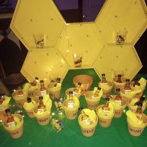 May include: A display featuring miniature Jack Daniel's Honey bottles in terracotta pots with the word "HUNNY" printed on them. The pots are arranged on a green surface, with a yellow honeycomb-shaped shelf in the background. Clear cups filled with candy are also present.