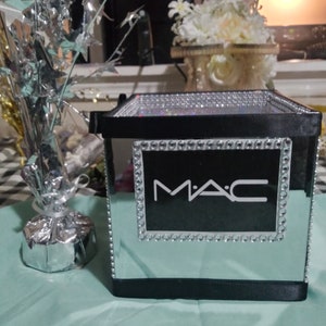 May include: A black mirrored box with a silver trim and the word "MAC" in white letters. The box is sitting on a light green surface.