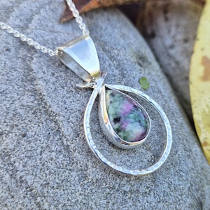 Op de afbeelding: Een zilveren hangerketting met een druppelvormige edelsteen met roze en groene tinten. De edelsteen is gezet in een gehamerd zilveren frame, hangend aan een fijne ketting. De hanger heeft een uniek, artistiek ontwerp.