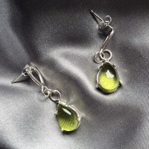 Pode incluir: Brincos de prata com pedras preciosas em forma de gota, verde-oliva. Os brincos têm um design curvo distinto, com um pequeno laço que conecta a gema à haste. As gemas são definidas em uma moldura de prata.