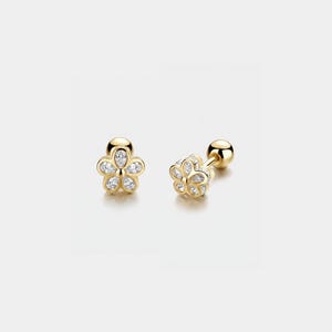 Puede incluir: Dos pendientes dorados con forma de flor, cada uno con pequeñas piedras preciosas transparentes engastadas en los pétalos. Los pendientes tienen cierre de poste y bola. Fondo claro.