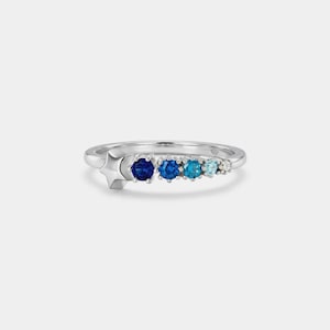 May include: A silver ring featuring a star and a row of blue gemstones. The gemstones graduate in color from dark blue to light blue. The ring has a simple, elegant design.