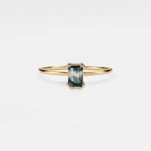 Puede incluir: Un anillo de oro con una gema rectangular verde musgo. El anillo tiene una banda delgada y delicada, y la gema está engastada en un engaste de cuatro puntas. La gema tiene un patrón natural único, sobre un fondo blanco.