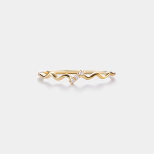 May include: A delicate gold ring with a wavy band design. The ring features three small, clear gemstones set in a diagonal line. The ring is set against a plain white background.