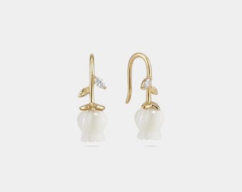 May include: A pair of gold-colored earrings featuring a white flower design. Each earring has a curved hook, a small leaf, and a clear gemstone accent. The flower is a delicate, bell-shaped design.
