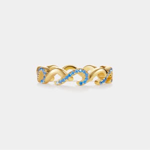 May include: A gold-colored ring with a repeating, intertwined design. The ring is adorned with small, light blue gemstones. The gemstones are set along the curves of the ring, adding a touch of sparkle. The ring is set against a plain white background.