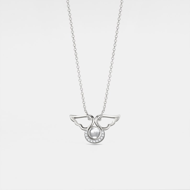 May include: A silver necklace with a pendant shaped like two angels with wings intertwined. The pendant has a small round pearl in the center and is surrounded by small clear stones.