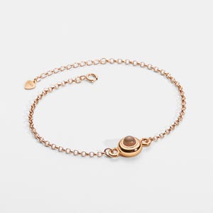 May include: Rose gold bracelet with a delicate chain and a central, round, light brown stone. The bracelet has a small heart-shaped tag near the clasp. The design is simple and elegant.