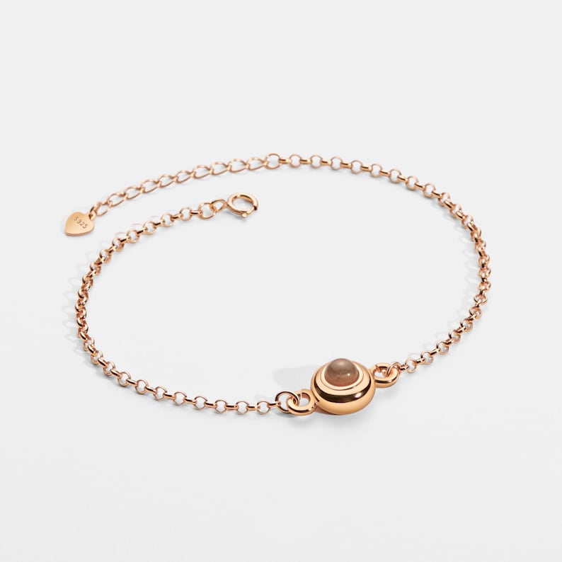 May include: Rose gold bracelet with a delicate chain and a central, round, light brown stone. The bracelet has a small heart-shaped tag near the clasp. The design is simple and elegant.