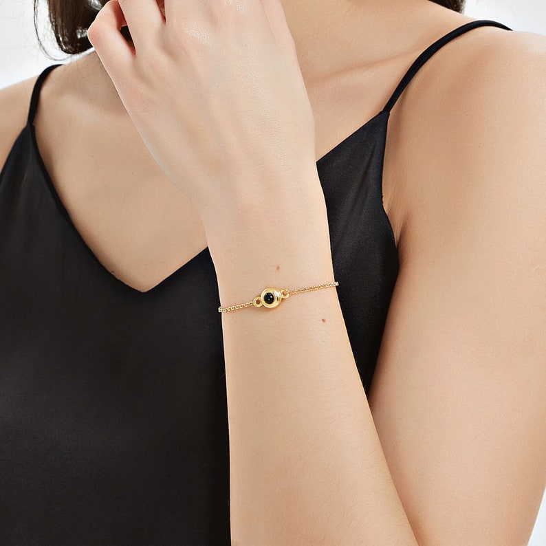 May include: A delicate gold bracelet featuring a small, round black gemstone at its center. The bracelet's chain is thin and elegant, with a simple clasp. The jewelry is worn on a wrist against a black top.