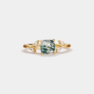 May include: A gold ring featuring a large, square-cut, moss agate gemstone with green and white inclusions. The ring is adorned with leaf-shaped accents and smaller, clear gemstones. The band has a delicate, nature-inspired design.