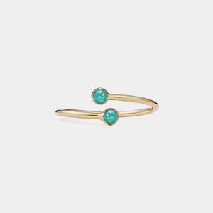 May include: A gold-colored ring with a unique, open design. The ring features two round, light blue opal stones. The band curves around, creating an elegant and modern look. The ring is set against a plain white background.