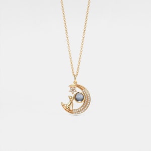May include: Gold necklace with a crescent moon pendant featuring a cat reaching for a star. The moon is set with clear stones and the star has a single clear stone in the centre.