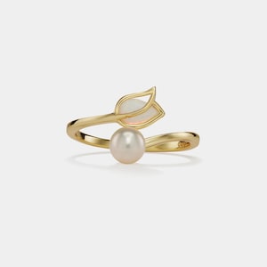 May include: A gold-colored ring featuring a freshwater pearl and a leaf-shaped design with a white gemstone. The ring's band curves around the finger, creating an elegant and nature-inspired aesthetic. The ring is set against a plain white background.