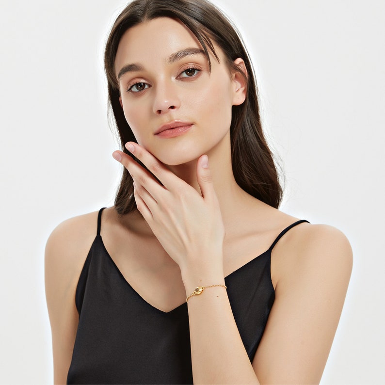 May include: A delicate gold bracelet with a small, round charm is worn on a wrist. The bracelet is on a slender chain and is set against a black top. The background is a plain white.