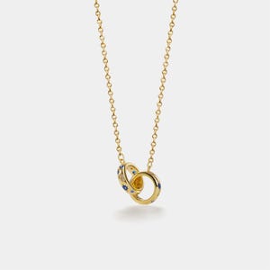 May include: A gold-colored necklace featuring two interlocking rings. The rings are adorned with small blue and clear gemstones. The necklace chain is also gold-colored and delicate, creating an elegant and sophisticated look.