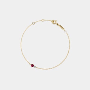 May include: Elegant gold bracelet with a rich red gemstone and a small, sparkling white stone. The bracelet has a fine chain and a secure clasp. A small tag with text is attached to the chain.