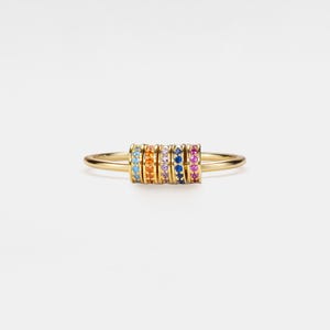 Puede incluir: Un anillo de oro con una serie de bandas de colores con gemas. El anillo muestra un espectro de colores, incluyendo azul, naranja, amarillo, morado y rosa, sobre un fondo blanco brillante. El diseño del anillo es simple pero llamativo.