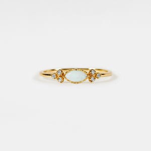 Könnte beinhalten: Ein zarter goldener Ring mit einem zentralen ovalen, hellblauen Edelstein, flankiert von Gruppen kleiner, funkelnder, klarer Steine. Der Ring hat ein dünnes, poliertes Band, das ein elegantes und minimalistisches Design schafft. Ein schönes Schmuckstück.