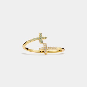 May include: A gold-colored ring with two cross designs. One cross is adorned with small, light green stones, and the other with clear, sparkling stones. The ring has a delicate, open design.