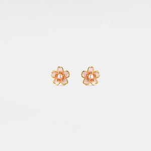 May include: A pair of flower-shaped stud earrings. Each earring features a pink enamel flower with a gold-colored outline and a small, round pearl in the center. The earrings are set against a white background.