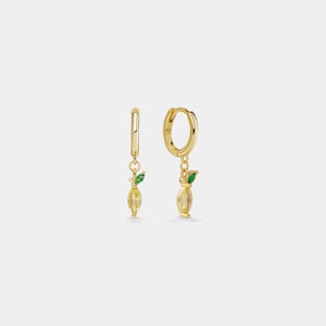 May include: A pair of gold hoop earrings featuring a dangling lemon-shaped charm. Each charm consists of a yellow marquise-cut crystal body and a small green marquise-cut crystal leaf accent. The hoops have a polished finish and a secure clasp.