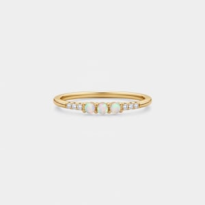 May include: A delicate gold ring featuring three opal stones and small, sparkling accent stones. The ring has a thin band and a minimalist design, perfect for everyday wear. The opal stones are round and have a milky white appearance.