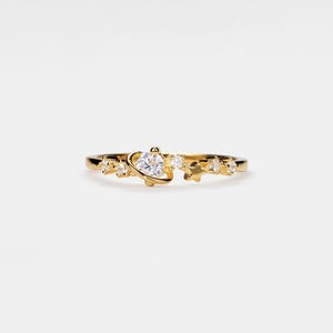 May include: A gold ring with a clear gemstone set in a small circle with a curved line around it, representing a planet. The ring is adorned with smaller clear gemstones, resembling stars.